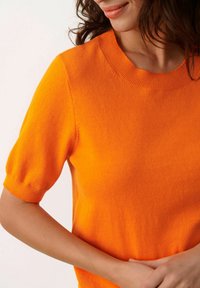 Kurzärmeliges Pullover mit Rundhalsausschnitt in leuchtendem Orange. Weiches Strickmaterial mit glatter Textur und dezent geripptem Kragen und Ärmeln.