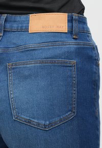 Μπλε τζιν παντελόνια με δερμάτινο λογότυπο στη μέση με ανάγλυφο το "NOISY MAY." Διαθέτουν δύο πίσω τσέπες και ραφές σε αντίθεση.