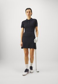 Černé golfové šaty s krátkým rukávem a polo límečkem, s vertikálním prošíváním, doplněné o bílé boty a šedou golfovou rukavici, držící golfovou hůl.