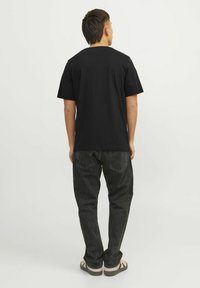 Schwarzes T-Shirt mit kurzen Ärmeln aus Baumwolle, mit lockerer Passform. Getragen mit dunklen Jeans und lässigen Sneakern, von hinten betrachtet.