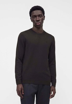 Jeune homme portant un pull sombre à manches longues et un pantalon sombre sur un fond clair uni, debout avec les bras détendus.