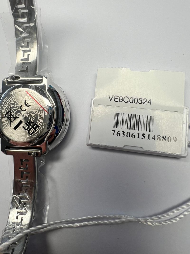 Srebrny metalowy zegarek na nadgarstek z okrągłą kopertą, wzorzystym paskiem i grawerowanym tyłem. Metka z code'ami kreskowymi i kodem produktu.