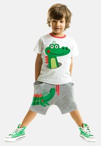 Strój dziecka składający się z białej koszulki z graficznym motywem zielonego krokodyla oraz szarych spodenek z podobnym wzorem. Zielone akcenty i czerwone detale.