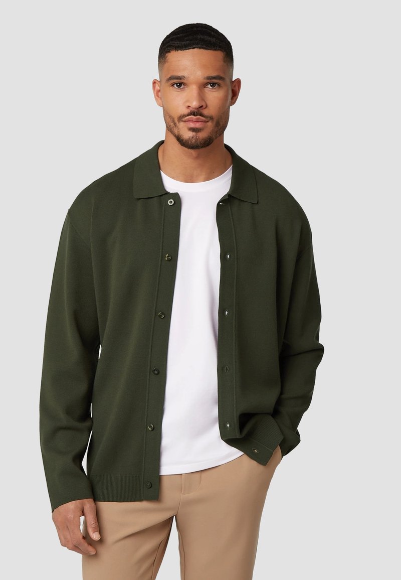 Donkergroene knoopkast cardigan met een zachte textuur, grote kraag en geribde manchetten, gedragen over een wit t-shirt en beige broek.