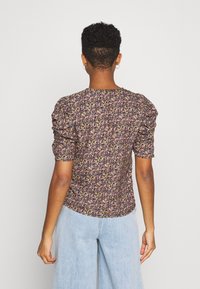 Femme avec des cheveux courts et bouclés portant un haut à manches bouffantes florales violettes et un jeans bleu clair, se tenant face à un fond blanc uni.