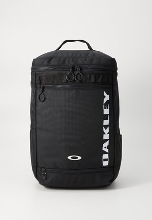 Sac à dos rectangulaire noir Oakley avec poignée supérieure, poches avant zippées et logo Oakley blanc vertical sur le côté droit.