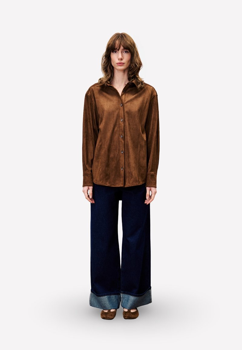 Brown suede button-up shirt with a pointed collar, paired with wide-leg dark denim jeans featuring a cuffed hem.