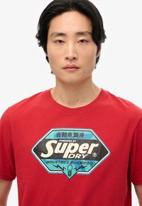 Camiseta roja de manga corta con un gran gráfico en azul y negro que presenta una forma de diamante y el texto "Impulsado por Super Dry Industries."