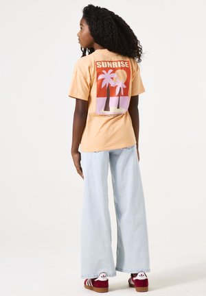 Perzikkleurig t-shirt met een palmboomafbeelding in rood, roze en wit. Gecombineerd met lichtblauwe wijde jeans en roodgestreepte schoenen.