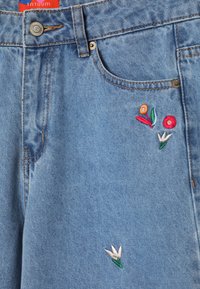 Jeans in denim blu chiaro con cuciture arancioni, due tasche frontali e ricami floreali con design rossi, arancioni e bianchi sul davanti.