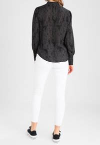 Blusa nera a maniche lunghe con un motivo sottile, colletto alto e maniche arricciate, abbinata a jeans skinny bianchi e sneakers nere.