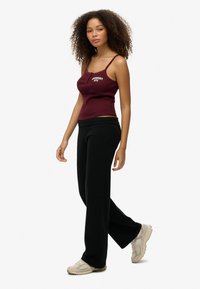 Bordeaux kleurige aansluitende tanktop met dunne bandjes en witte tekst, gecombineerd met zwarte flared trainingsbroek en beige sneakers, op een model.