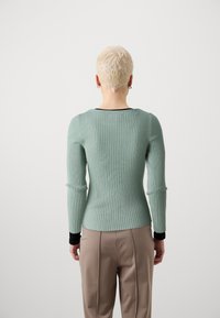 Ribattu, pitkähihaineen mintunvihreä neule, joka on tiukasti istuva. Mustat yksityiskohdat hihoissa. Yllään beiget housut, kuvattu takaa.