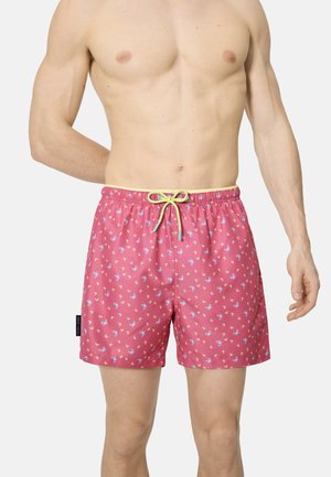 Hombre vistiendo pantalones cortos de baño rosa con pequeño estampado floral azul y amarillo, y cordón amarillo, sobre un fondo liso.