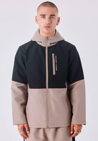 Chaqueta con capucha negra y beige con un bolsillo con cremallera, fabricada en un tejido texturizado. Presenta un diseño de bloques de color y un alto collar.