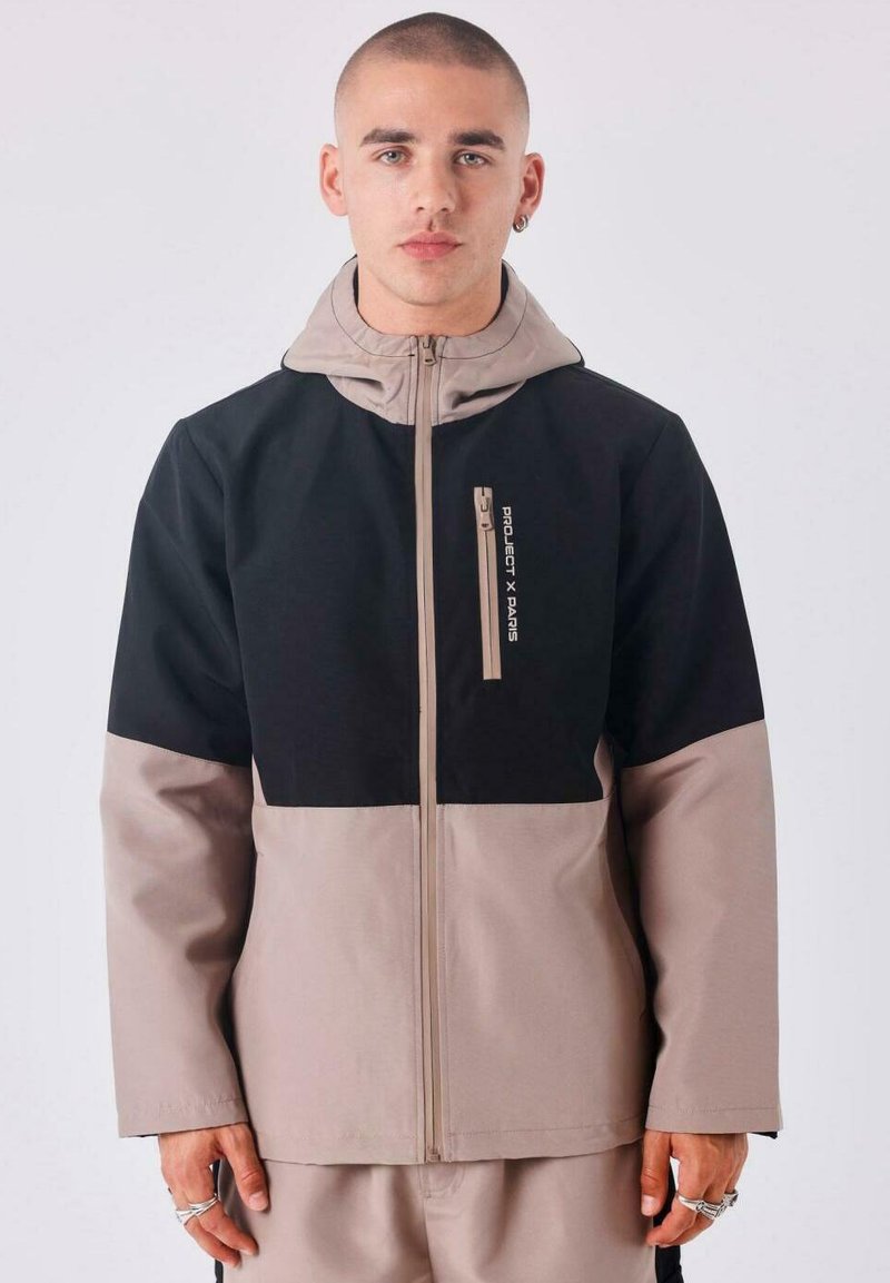Chaqueta con capucha negra y beige con un bolsillo con cremallera, fabricada en un tejido texturizado. Presenta un diseño de bloques de color y un alto collar.