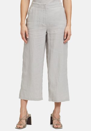 Kobieta ubrana w jasnoszare, szerokie spodnie o kroju capri oraz beżowe sandały na wysokim obcasie z paskami, stojąca na tle jednolitego tła.