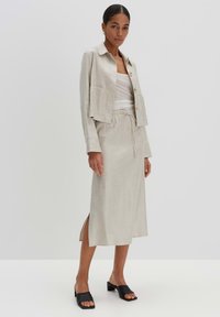 Beige linnen cropped jas met knoopsluiting, gecombineerd met een bijpassende midi-rok met een zijsplit. Zwarte instap sandaaltjes maken de outfit compleet.