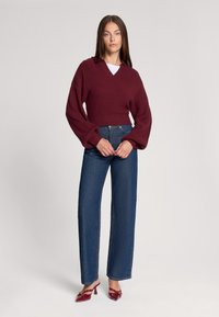 Burgundowy krótki sweter dzianinowy z dekoltem w serek i szerokimi rękawami, w zestawie z jasnoniebieskimi dżinsami z wysokim stanem oraz czerwonymi klapkami na obcasie.