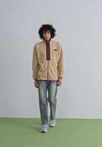 Beige fleece jacket with brown accents and zipper, paired with light blue jeans and gray sneakers, set against a plain background.