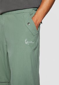 Shorts vert clair en tissu texturé, dotés d'une poche latérale avec un bouton-pression en métal, et d'un logo blanc brodé à l'avant.