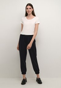 Witte katoenen t-shirt met korte mouwen, gecombineerd met marineblauwe joggingbroeken met een elastische tailleband en boordjes aan de enkels. Gedragen met zwarte instapper schoenen.