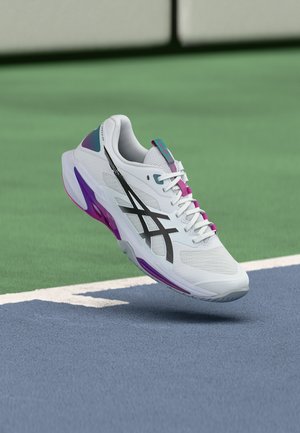 Λευκό αθλητικό παπούτσι με μαύρο λογότυπο και μωβ λεπτομέρειες που αιωρείται πάνω από την επιφάνεια αθλητικού γηπέδου σε μπλε και πράσινες αποχρώσεις.