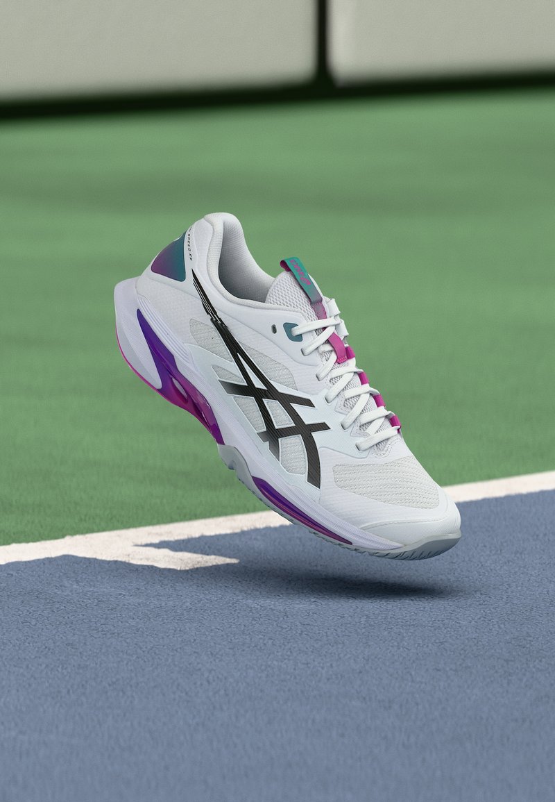 Λευκό αθλητικό παπούτσι με μαύρο λογότυπο και μωβ λεπτομέρειες που αιωρείται πάνω από την επιφάνεια αθλητικού γηπέδου σε μπλε και πράσινες αποχρώσεις.