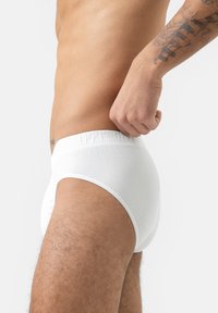 Culottes en coton blanc, taille élastique, design ajusté, texture lisse et couverture modérée. Présentées sur une silhouette masculine de côté.