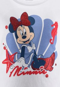 Gráfico de Minnie Mouse en una camiseta blanca, con un lazo rojo brillante, vestido azul y acentos de estrella de mar y caballito de mar en rojo y azul.