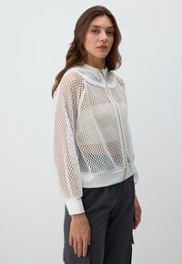 Felpa con zip bianca in mesh caratterizzata da un design leggero e testurizzato, con cappuccio e polsini e orlo bianchi. Dettaglio della zip dorata visibile.