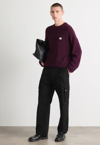 Maroon knit sweater with ribbed texture, paired with black cargo pants featuring side pockets and silver hardware. Holding a black bag.