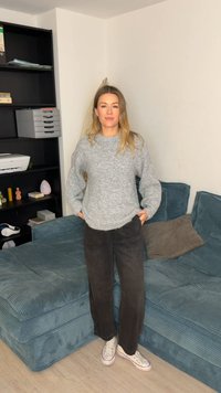 Grijze gebreide trui met losse pasvorm, zwarte corduroy broek en witte sneakers. Op de achtergrond een tealkleurige corduroy bank en planken.