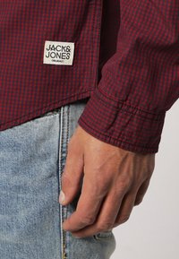 Main d'un homme à côté de la poche d'un jean bleu portant une chemise à carreaux rouge et noir avec l'étiquette Jack & Jones Originals visible.