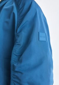 Chaqueta bomber azul con una textura suave, presenta costuras fruncidas, un discreto parche con el logo en la manga y un corte estructurado.