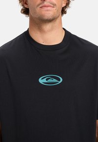 Camiseta de algodón negra con cuello redondo y un logo de ola turquesa en el pecho. Textura suave con mangas cortas.