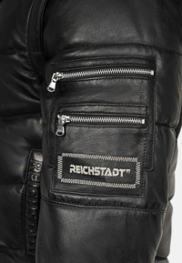 Zwarte leren pufferjack met twee ritszakken, een logo-opdruk met witte stiksels en een gestructureerde afwerking.