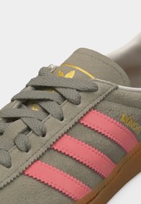 Γκρι σουέτ αθλητικό παπούτσι με ροζ ζιγκ-ζαγκ ρίγες, γκρι κορδόνια, χρυσό λογότυπο στη γλώσσα και σόλα καραμέλας, κοντινή λήψη στις πλευρικές και πάνω λεπτομέρειες.