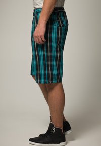 Pantaloncini a quadri in nero, turchese e grigio; con tasca laterale, design fino al ginocchio e tessuto leggero. Indossati con sneakers nere.