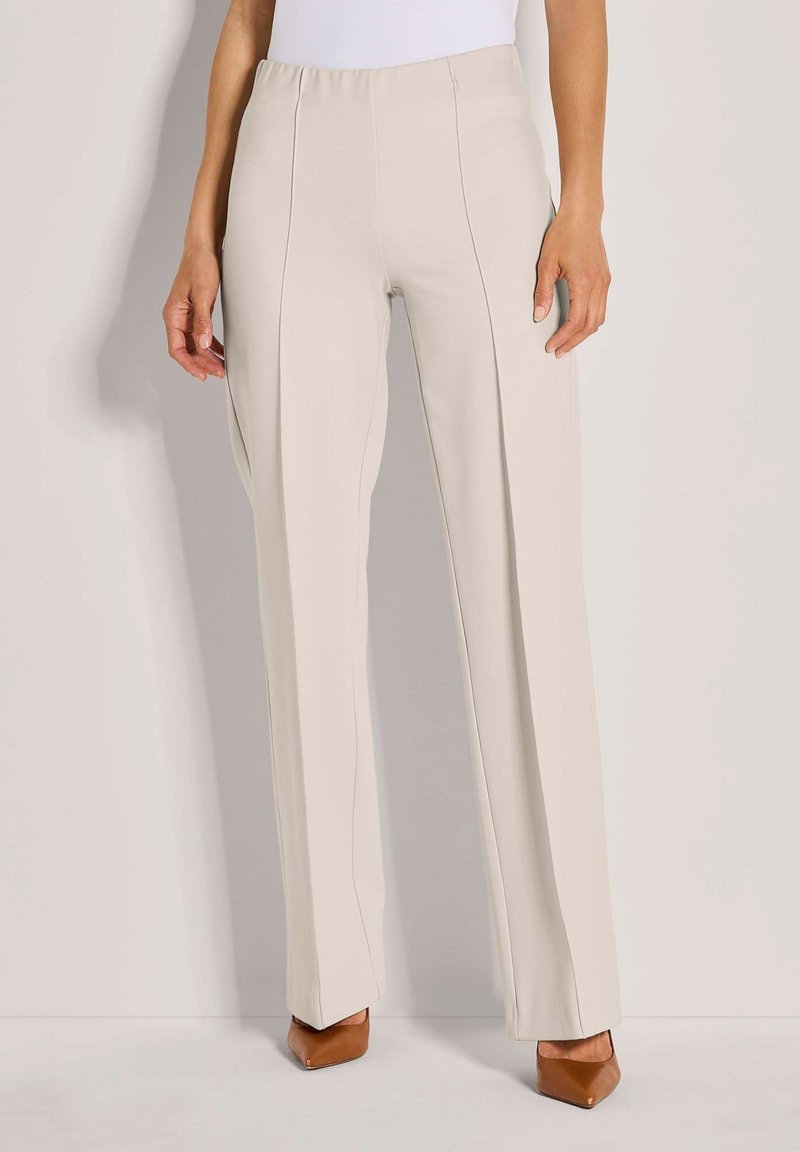 Beige hoge taille rechte pijpen broek met voornaad, gedragen met bruine spitse schoenen en een witte top die zichtbaar is bij de taille.