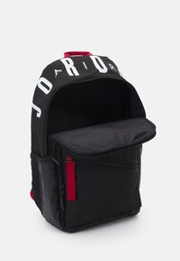 Rucsac negru cu logo alb mare, fermoar roșu, buzunar frontal și compartiment principal spațios. Materialul pare a fi o țesătură durabilă.