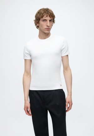 Νέος άνδρας με καστανά σγουρά μαλλιά φοράει στενό λευκό κοντομάνικο πουκάμισο και άνετο μαύρο παντελόνι, στέκεται μπροστά σε απλό ανοιχτό φόντο.