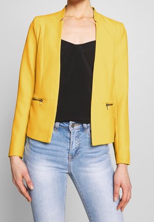 Kvinde iført en strålende gul blazer med lynlåslommer, sort top og lyseblå falmede jeans, stående foran en ensfarvet baggrund.