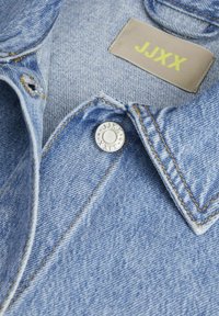 Šviesiai mėlynas džinsinis švarkas su smailiu apykakle, geltona etikete su "JUXX" ir sidabriniais mygtukų detalėmis. Išskirtinis siuvimo akcentai visame švarke.