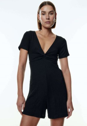 Combinaison noire à manches courtes avec un décolleté en V et un détail à l'avant en torsion. Fabriquée en tissu texturé, elle présente une silhouette ajustée et une coupe short.