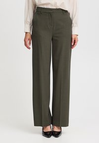 Olivgrüne maßgeschneiderte Hose mit weitem Bein, strukturiertem Stoff, vorderen Falten und Gürtelschlaufen, kombiniert mit schwarzen High Heels.