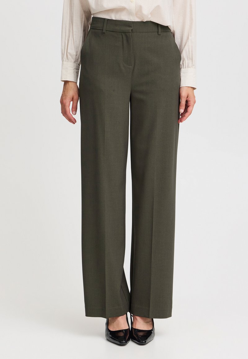 Olivgrüne maßgeschneiderte Hose mit weitem Bein, strukturiertem Stoff, vorderen Falten und Gürtelschlaufen, kombiniert mit schwarzen High Heels.