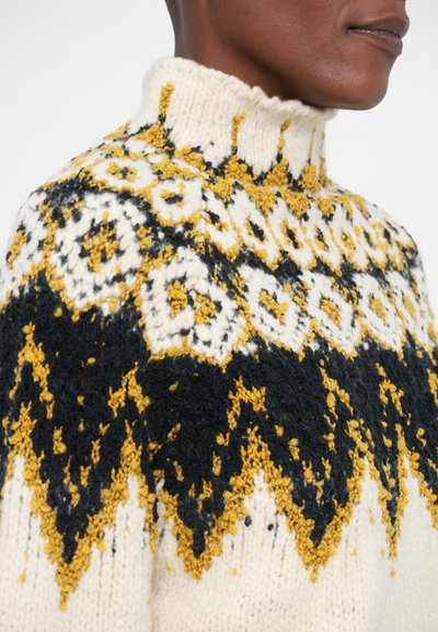 Primo piano di una persona che indossa un maglione a collo alto lavorato a maglia con motivi geometrici neri, gialli e bianchi.