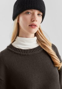 Donkergrijze gebreide trui met een ronde hals, gedragen over een witte coltrui. Het model heeft lang blond haar en draagt een zwarte geribbelde beanie.