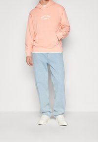 Helle pinke Kapuzenjacke mit "PORTLAND" Logo, vorderer Tasche und gerippten Bündchen, kombiniert mit hellblauen Jeans und weißen Sneakers.
